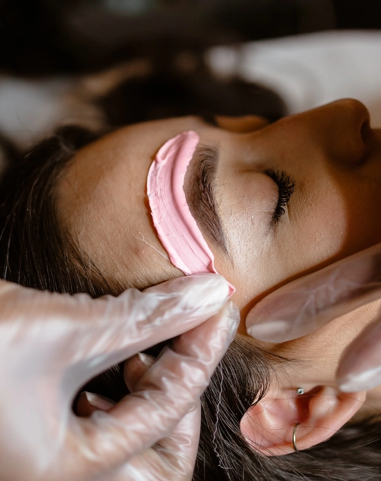 Facial skincare treatment in progress with a pink mask being applied to a woman's cheek.
